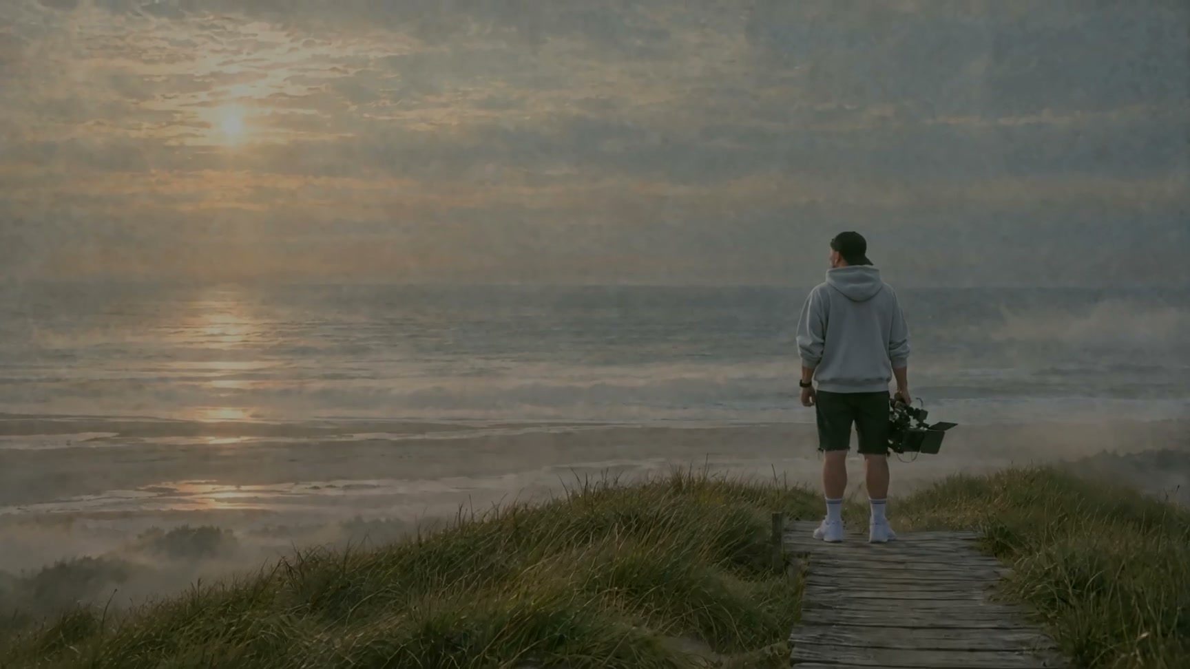Leon Saß im grauen Hoodie steht von hinten gesehen mit seiner Kamera auf einem Holzsteg in den Dünen und blickt auf die Nordsee