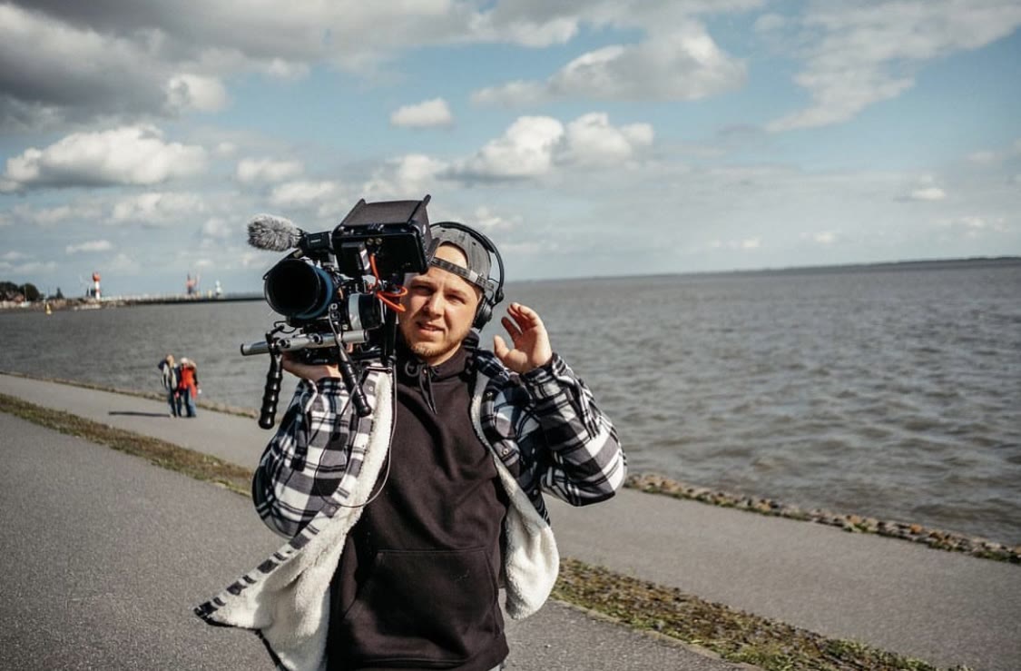 Leon Saß mit Kopfhörern und Filmkamera auf der Schulter auf einem Deich am Wasser
