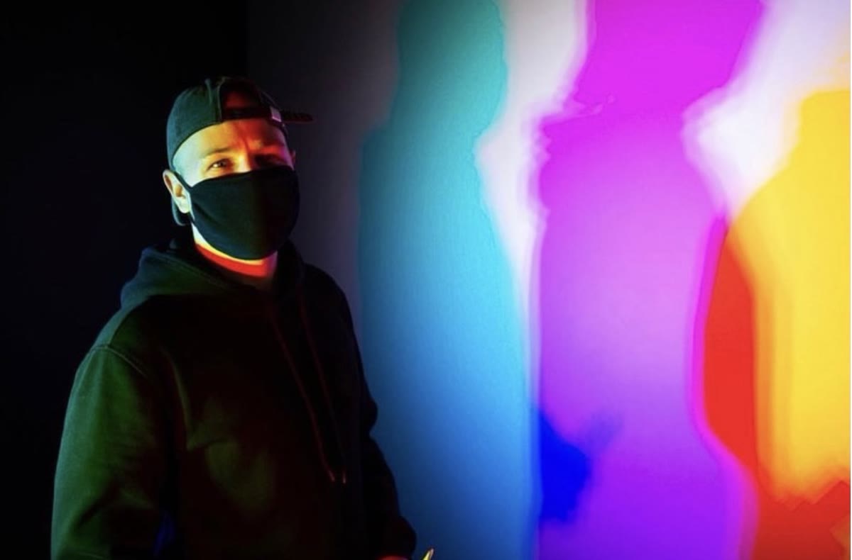 Leon Saß mit Cap und schwarzer Maske vor einer farbigen Lichtprojektion in Türkis, Magenta und Gelb