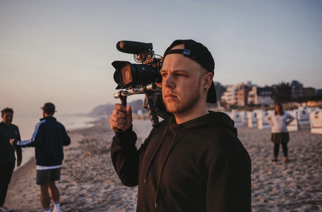 Leon Saß mit Kamera-Rig auf der Schulter bei Sonnenaufgang an einem Strand mit Strandkörben, im Hintergrund sein Team