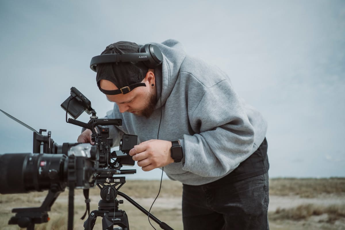 Leon Saß mit Kopfhörern im hellgrauen Hoodie beugt sich über seine Filmkamera auf dem Stativ, im Hintergrund Dünen